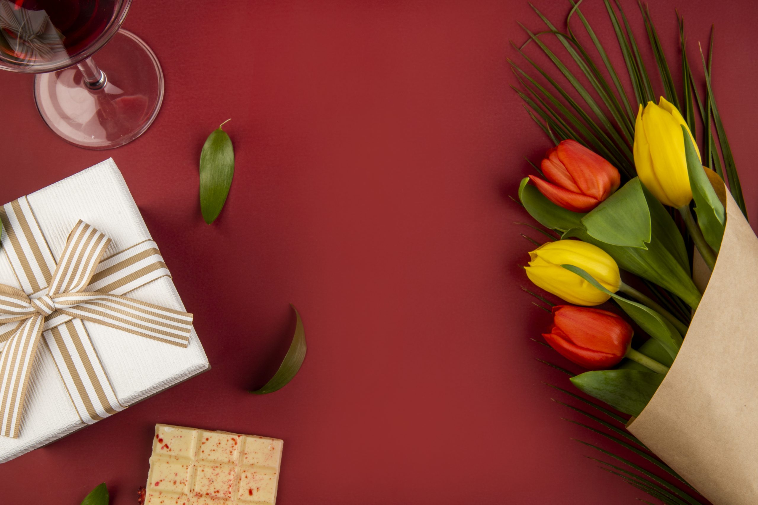 top view of a bouquet of red and yellow color tulips with a glass of wine, white chocolate and a gift box on red background with copy space
